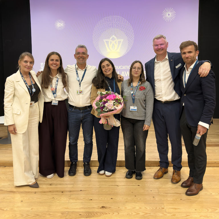 Group of seven adults on a stage, posing for a photo; one person holds a bouquet of pink flowers, all wearing event lanyards against a purple backdrop with a logo and text.