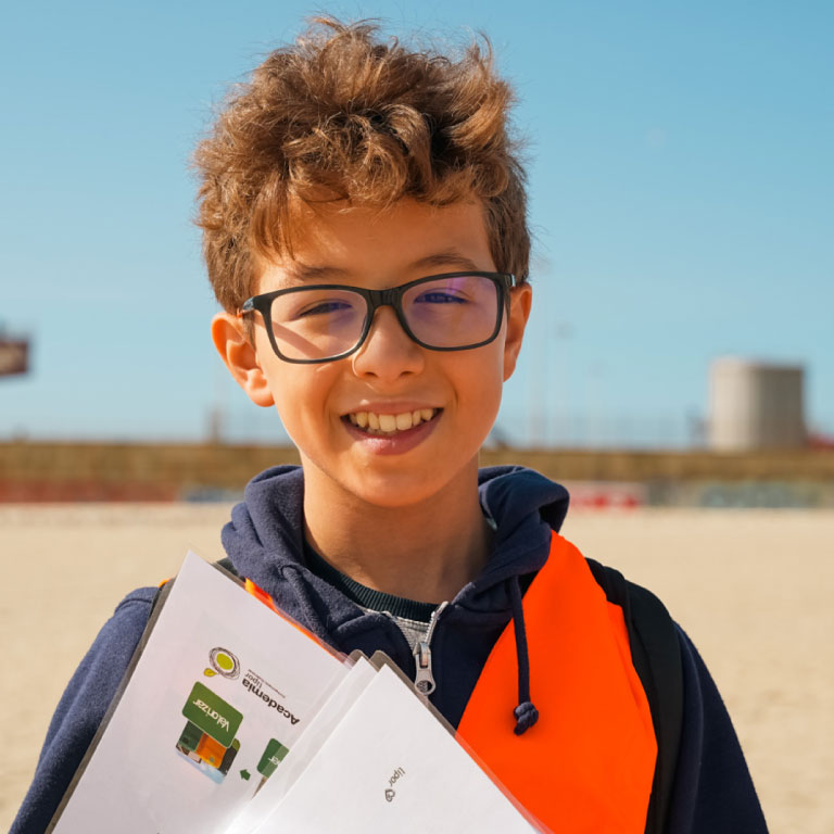 Young boy wearing a bright orange life vest and glasses, smiling at the beach while holding papers or worksheets close to his chest.